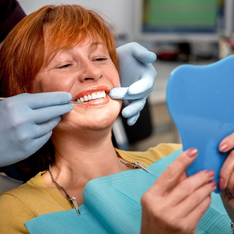 Satisfied senior woman looking his teeth in a mirror after getting dentures at dentist's office.